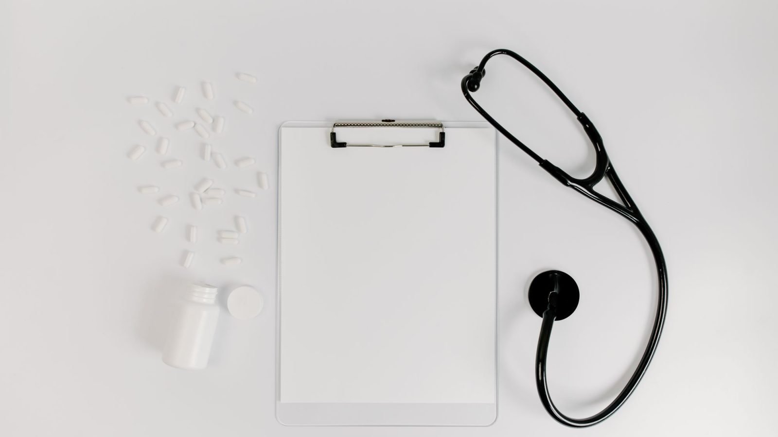 A clipboard with paper beside a stethoscope and pills