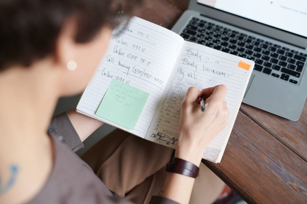 A person writing in a notebook next to a laptop