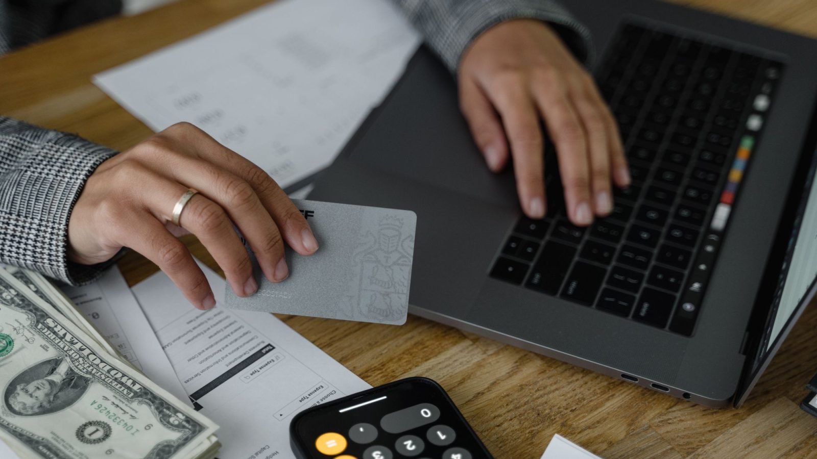 A hand holding a credit card while using a laptop, with a calculator and cash nearby