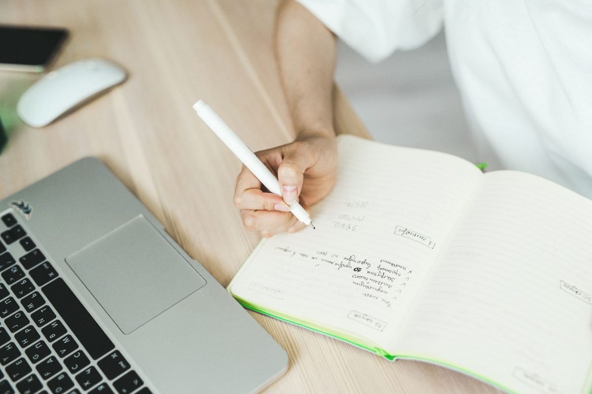 A hand writing in a notebook near a laptop