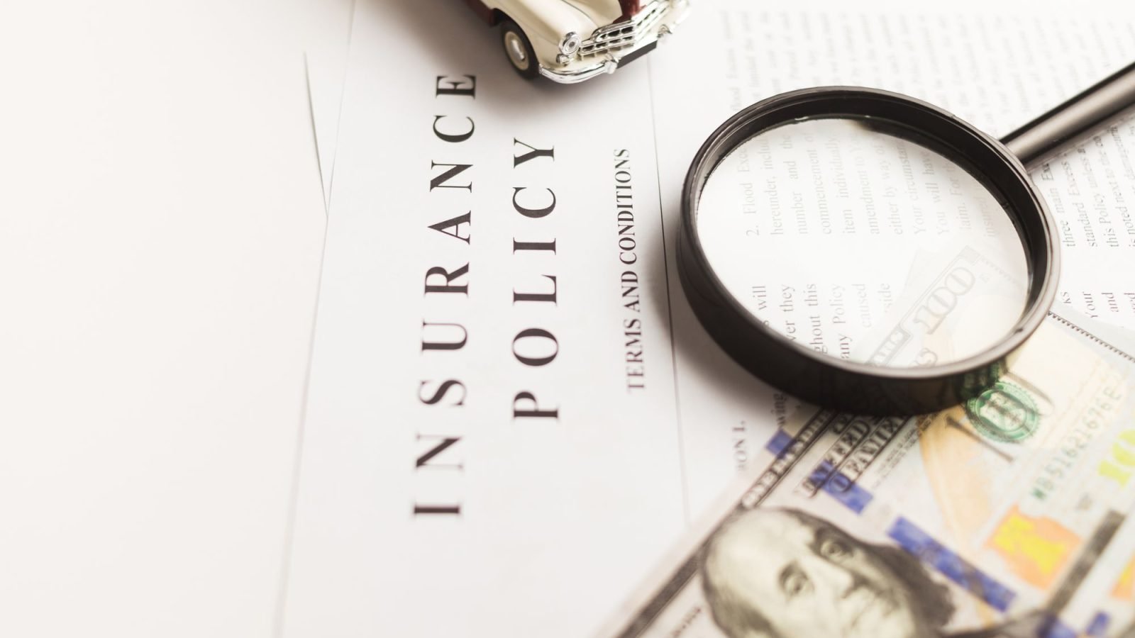 An insurance policy document with a magnifying glass, a toy car and money on a desk