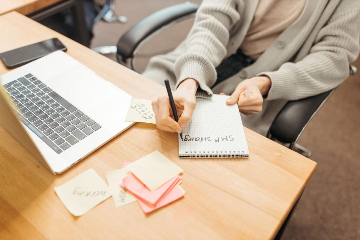 An adult writing notes in a notebook at a desk with a laptop