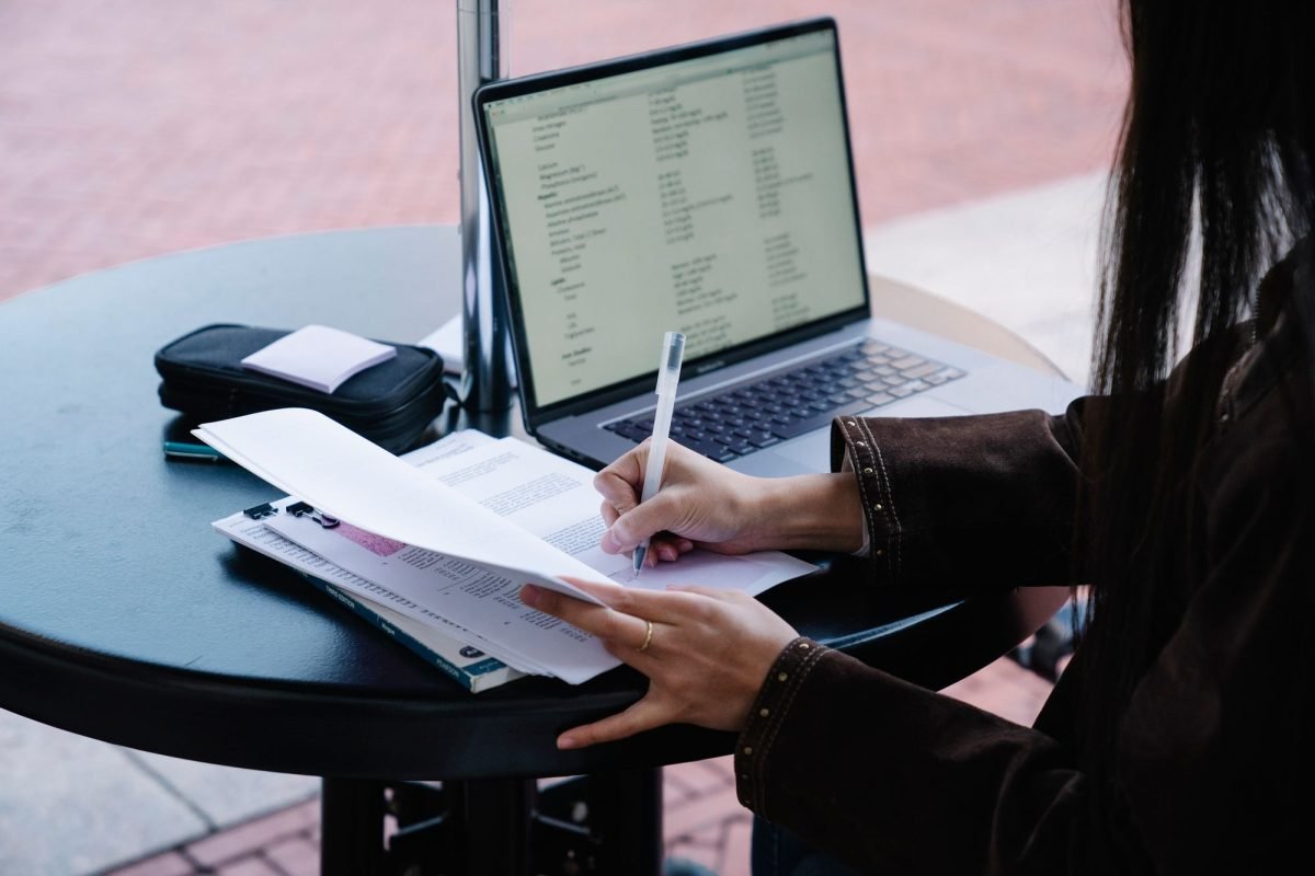 A woman writing on papers at an outdoor cafe table with a laptop