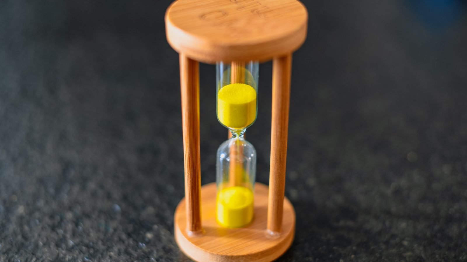 A wooden hourglass with yellow sand on a dark textured surface