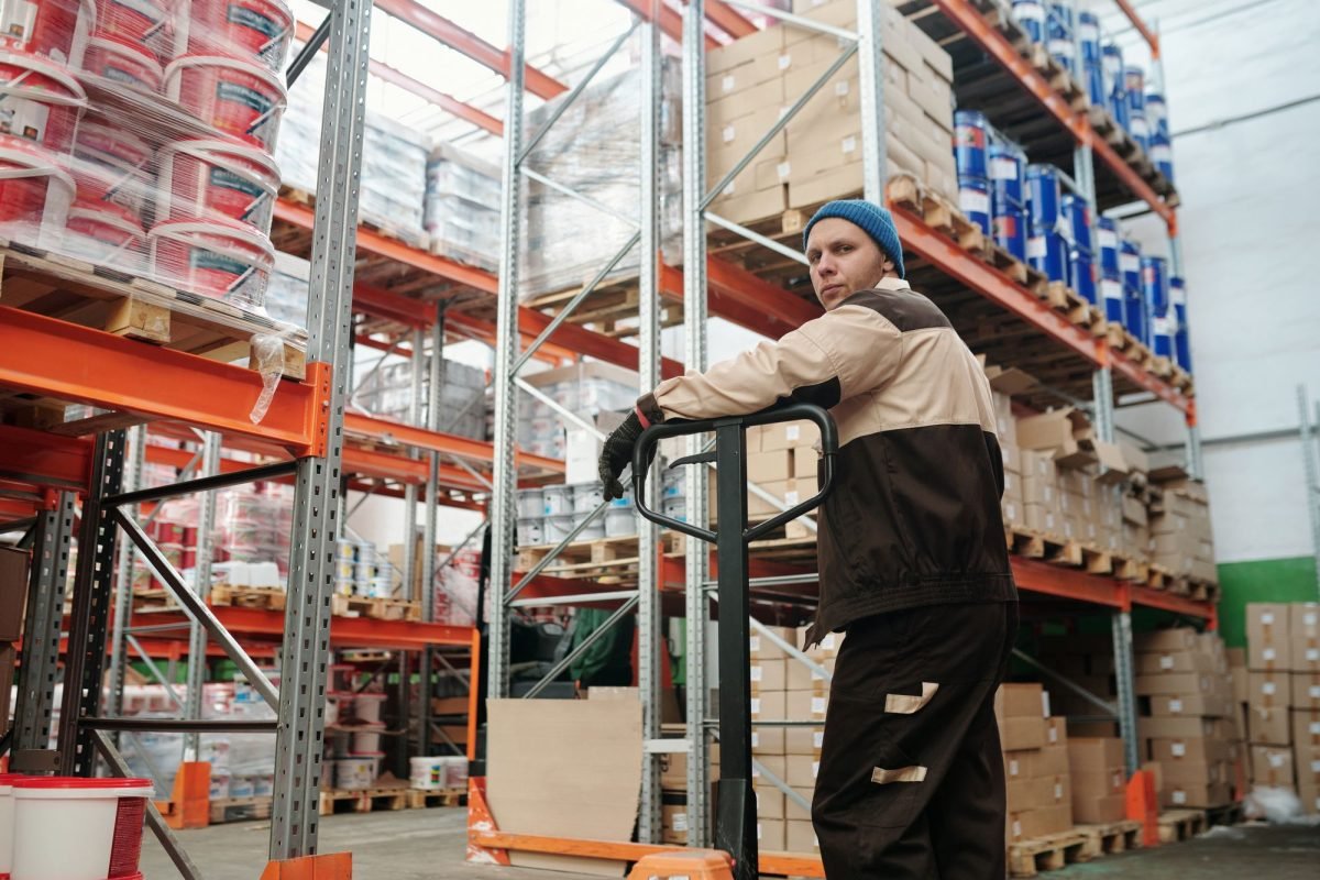 A warehouse worker operating a pallet jack among stacked boxes